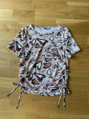 Hjärtan topp från FB Sister med snörning - Kortärmad topp från FB Sister med hjärtmönster i beige, brun och vit. Toppen har snörning i sidorna för en snygg detalj och är tillverkad i ett mjukt, stretchigt material. Perfekt för dig som gillar unika prints och trendiga detaljer.