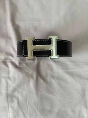 Svart bälte med stor H-spänne - Snyggt svart bälte i skinn med ett stort, silverfärgat H-format spänne. Bältet har vita kontrastsömmar längs kanterna som ger en clean och modern look. Perfekt accessoar för att lyfta vilken outfit som helst.
