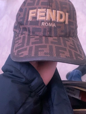 Brun Fendi keps med logomönster - Säljer en brun keps från Fendi med det klassiska FF-logomönstret över hela kepsen och broderad FENDI ROMA-text framtill. Kepsen har böjd skärm och justerbar baksida. Materialet är troligtvis bomull eller en bomullsblandning. Perfekt för dig som gillar lyxiga accessoarer.