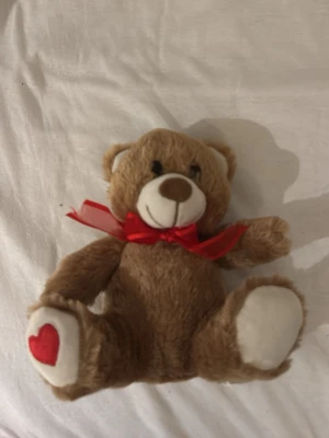 Brun nallebjörn med röd rosett - Söt mjuk nallebjörn i ljusbrunt plyschmaterial med vit nos och tassar. Den har en röd rosett runt halsen och ett rött hjärta broderat på ena tassen. Perfekt som present eller dekoration.