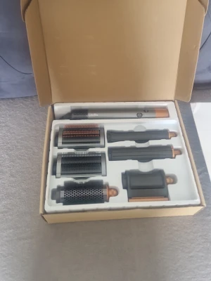 Dyson Airwrap multistyler set - Säljer ett Dyson Airwrap multistyler set i grå och kopparfärg. Setet innehåller flera olika stylingtillbehör för hår, inklusive borstar och locktillbehör. Allt ligger i originalförpackning och är perfekt för dig som vill fixa olika frisyrer hemma.