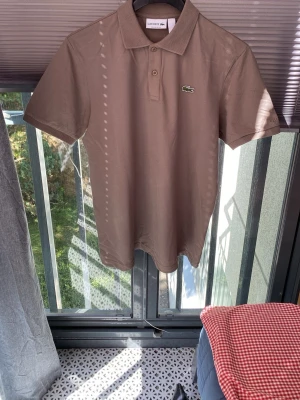 Beige pikétröja från Lacoste - Säljer en klassisk beige pikétröja från Lacoste med korta ärmar och krage. Tröjan har knappar framtill och den ikoniska gröna krokodilloggan på bröstet. Perfekt till sommaren och enkel att matcha med olika stilar.