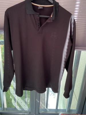 Svart långärmad piké från BOSS - Stilren svart långärmad pikétröja från BOSS med klassisk krage och knäppning framtill. Diskret BOSS-logga på bröstet. Perfekt till både jeans och chinos för en clean look.