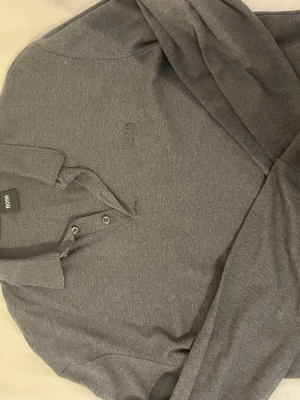 Grå långärmad pikétröja från HUGO BOSS - Snygg grå långärmad pikétröja från BOSS med klassisk krage och två knappar framtill. Diskret BOSS-logga broderad på bröstet. Tillverkad i mjukt och bekvämt material som passar perfekt till en avslappnad men stilren look.