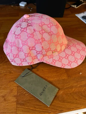 Rosa Gucci keps med GG-mönster - Säljer en snygg rosa keps Gucci med det klassiska GG-monogrammet i hela tyget. Kepsen har böjd skärm och är tillverkad i ett slitstarkt tyg med en liten guldfärgad detalj på sidan. Perfekt för dig som vill sticka ut med en exklusiv accessoar.