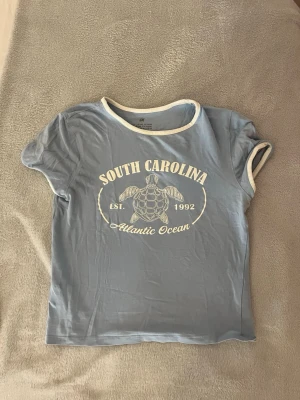 Blå t-shirt med South Carolina-tryck - Snygg ljusblå t-shirt med vit kant vid hals och ärmar samt tryck med texten 'South Carolina Atlantic Ocean' och en sköldpadda på bröstet.