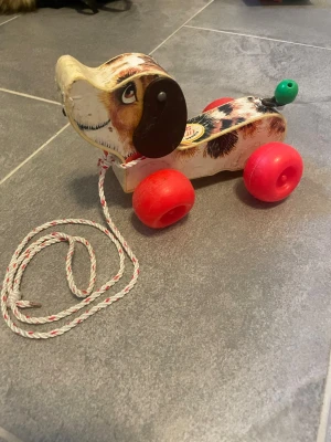 Fisher Price dragleksak hund - Säljer en klassisk dragleksak i form av en hund från Fisher Price, modell Little Snoopy. Leksaken är gjord i trä med tryckt motiv, har röda hjul, svart öron och en grön svans. Dragsnöre i vitt och rött medföljer. Perfekt för yngre barn som gillar att dra runt sina leksaker.
