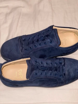   Marinblå Christian Louboutin - Snygga mörkblå sneakers i mocka med matchande blå skosnören och ikonisk röd sula med signaturdetalj. Insidan är ljusbeige och skon har en klassisk låg profil som passar till många olika stilar.