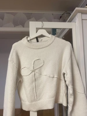 Vit stickad tröja med rosett-detalj - Säljer en krämvit stickad tröja med rund hals och ribbade muddar. Tröjan har en unik rosett-detalj framtill som ger en söt och trendig look. Perfekt för dig som gillar stilrena plagg med en twist. Materialet känns mjukt och mysigt.