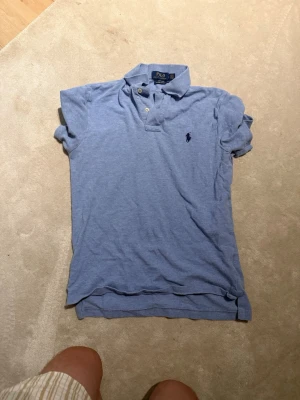 Blå pikétröja från Ralph Lauren - Snygg ljusblå pikétröja från Ralph Lauren med klassisk krage och knappar framtill. Tröjan har broderad logga på bröstet och är i en slim fit-modell. Perfekt för en stilren och avslappnad look.