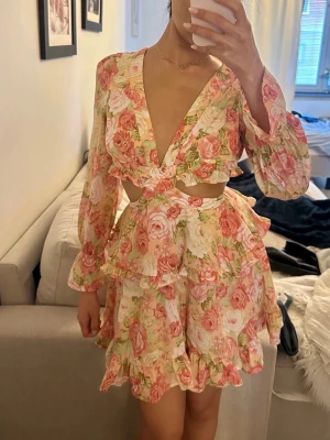 Blommig kortklänning med volanger - Supersöt kortklänning med blommigt mönster i rosa, vitt och grönt. Klänningen har djup v-ringning, långa ärmar med volangkant, cut-out detaljer i midjan och omlottknytning i ryggen.  Oanvänd! 