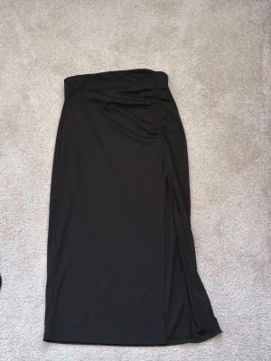Svart långkjol med slits - Säljer en svart långkjol med hög midja och snygg slits på sidan. Kjolen har en lätt rynkad detalj i midjan som ger en cool touch. Tillverkad i ett mjukt och stretchigt material som sitter bekvämt och följer kroppen.