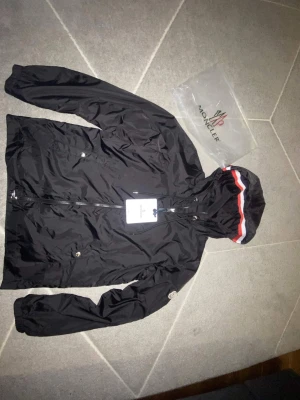 Svart vindjacka med huva från Moncler - Snygg svart vindjacka från Moncler med huva och dragkedja. Jackan har Monclers logga på ärmen och en vit-röd-blå rand på huvan. Tillverkad i lätt, glansigt material som står emot vind. Perfekt för dig som vill ha en stilren och sportig look. Size S.
