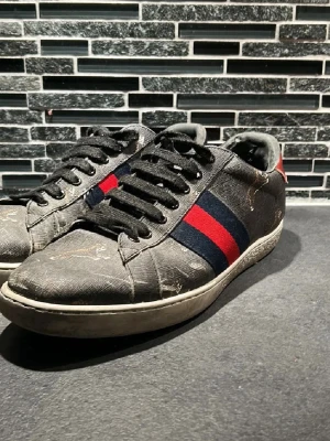 Svarta sneakers med röd/blå rand - Snygga svarta sneakers med klassisk röd och marinblå rand på sidan. Skorna har svarta skosnören och vit sula. Materialet ser ut att vara canvas med läderdetaljer och en röd detalj på hälen. Perfekta för dig som gillar streetstyle.