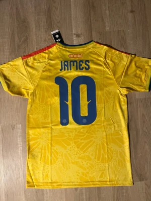 Gul Colombia matchtröja Rodriguez #10 - Snygg gul Colombia matchtröja med blå detaljer och röda ränder på axlarna. Nummer 10 och namnet James på ryggen. Broderad landslagssköld på bröstet. Tillverkad i lätt och ventilerande material, perfekt för fotboll. HELT NY!