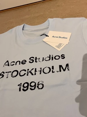 Ljusblå Acne Studios t-shirt - Säljer en ljusblå t-shirt från Acne Studios med svart tryck framtill där det står 'Acne Studios STOCKHOLM 1996'. Klassisk passform och rund halsringning. Perfekt för dig som gillar stilrena plagg med en twist.