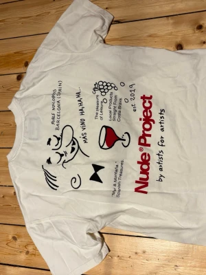 Creme vit t-shirt från Nude Project - Creme vit t-shirt från Nude Project med tryck på både fram- och baksida. Baksidan har ett stort grafiskt tryck med text och illustrationer i svart och rött. Framsidan har en mindre logga. Klassisk passform och rund hals. Perfekt för dig som gillar streetwear och unika prints. Min personliga favvotröja! Riktigt coola tryck på ryggen och en lite Boxig passform😍