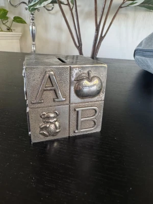Sparbössa kub i metall med bokstäver och djurmotiv - Spargris formad som en ABC-kloss. Varje sida har bokstäver och djurmotiv. Spargrisen har en öppning för mynt på ovansidan och en öppning på undersidan för att ta ut pengar. Som dopgåva.