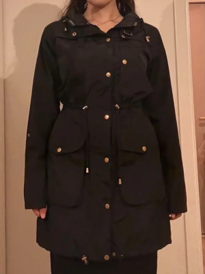 Regn/vind-kappa - Elegant och stilig regnkappa. Vindtät och vattnet, och håller verkligen värmen inne trots att den är tunn. Gå att justera vid midjan och längst ner, samt på luvan. Knappt använd.