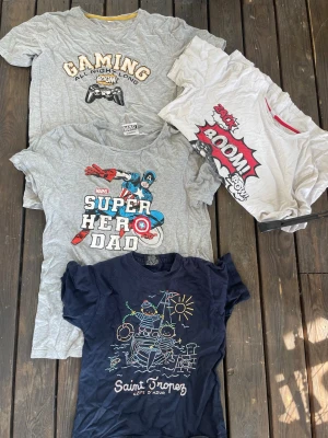 4-pack t-shirts gaming, Marvel & print - Fyra t-shirts i olika färger och tryck: en grå med gaming-tryck, en vit med serietidningsinspirerat BOOM-tryck, en grå Marvel Super Hero Dad och en marinblå med Saint Tropez-tryck från Spirit. Alla är kortärmade och i mjuk bomull, perfekta för en avslappnad stil.