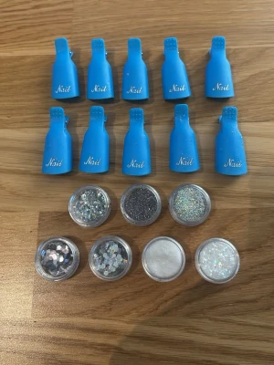 Blå nagelklämmor & glittermix - Säljer ett set med 10 nagelklämmor samt 7 burkar med olika glitter och paljetter i silver, vitt och olika former. Glittret är aldrig använt och klämmorna kommer att göras rent innan de har sålts. Allt är i bra skick.