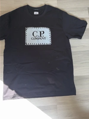 Svart C.P. Company t-shirt - Snygg svart t-shirt från C.P. Company med ett stort vitt patch-liknande tryck på bröstet och tydlig logga. Klassisk rund hals och korta ärmar. Perfekt till jeans eller shorts för en clean och modern look.