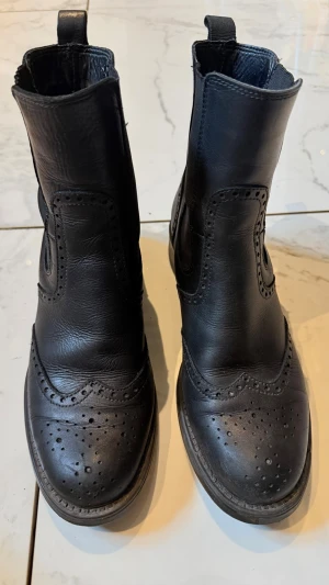 Svarta Chelsea boots med broguemönster - Säljer ett par svarta Chelsea boots i skinn med klassiskt broguemönster och rundad tå. Bootsens ovandel har dekorativa perforeringar och elastiska paneler på sidorna för enkel påtagning. Perfekta för dig som gillar stilrena och tidlösa skor.