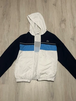 Lacoste vindjacka med huva - Snygg vindjacka från Lacoste i vitt, blått och marinblått. Jackan har huva, dragkedja framtill, elastiska muddar och två fickor. Klassisk Lacoste-logga på bröstet och sportig design med färgblock. Perfekt för en streetwear-look. Tyvärr ett litet hål som syns på bild 2 därav det bra priset! Förutom det är den i helt perfekt skick. Matchande byxor finns även i helt perfekt skick skriv vid intresse!!