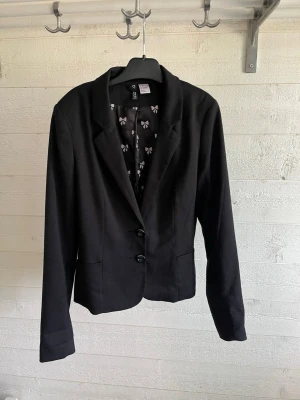 Svart kavaj från H&M, strl 34 - Stilren svart kavaj från H&M med två knappar framtill och figurnära passform. Fodret har ett gulligt mönster med små rosetter. Perfekt för att lyfta din outfit med en klassisk touch. Materialet känns mjukt och bekvämt.