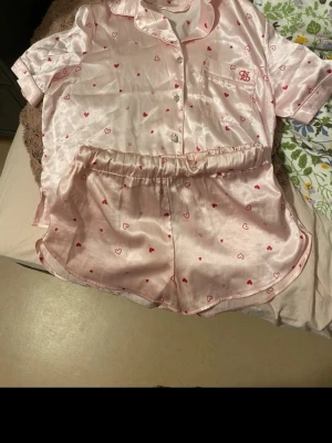 Rosa satinpyjamas med hjärtmönster - Söt pyjamas i glansigt rosa satin med kortärmad skjorta och matchande shorts. Setet har små röda och rosa hjärtan över hela tyget och en bröstficka med broderad detalj. Perfekt för mysiga kvällar hemma.