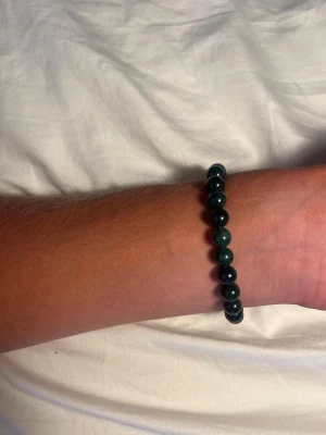 Mörkgrönt pärlarmband - Snyggt armband med mörkgröna, runda pärlor på elastisk tråd. Pärlorna har en glansig yta och varierar något i nyans från djupgrönt till blågrönt. Perfekt accessoar för att lyfta din stil med en enkel men trendig detalj.