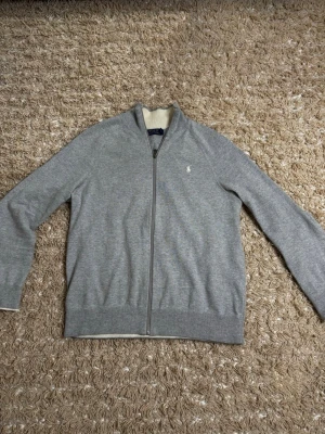 Grå stickad ziptröja Polo Ralph Lauren - Snygg grå stickad ziptröja från Polo Ralph Lauren med diskret logga på bröstet. Tröjan har hel dragkedja, ribbade muddar och en krage med vit insida. Perfekt till lager på lager och enkel att matcha. Fits Small