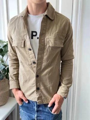 Calliope overshirt  - Stilren beige overshirt med knappar framtill och två bröstfickor med lock. Modellen är 180cm och väger 75kg, rekommenderas inte för folk som är större än det. Perfekt att bära öppen över en t-shirt för en avslappnad look.