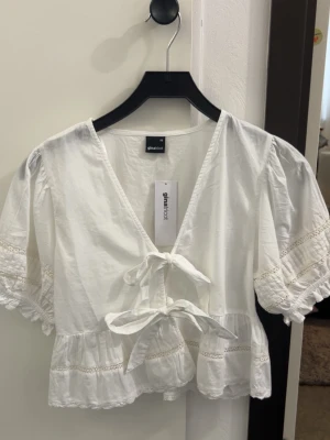 Vit blus med knyt från Gina Tricot - Söt vit blus från Gina Tricot med v-ringning, puffiga korta ärmar och två knyt framtill. Blusen har volangkant nedtill och spetsdetaljer längs ärmar och fåll. Perfekt för en trendig och luftig look. Helt ny 