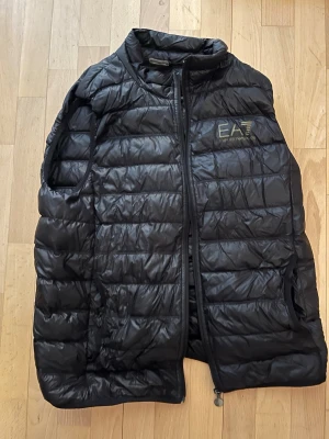 Svart dunväst från Emporio Armani - Snygg svart dunväst från Emporio Armani EA7 med quiltad design och hög krage. Västen har dragkedja framtill och diskret logga på bröstet. Perfekt för lager på lager och ger en sportig, modern look. Och om du vill prata om pris det är bara skriva till mig