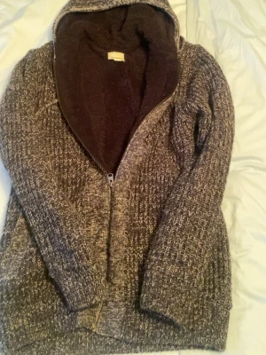 Brunmelerad stickad hoodie med dragkedja - Mysig stickad hoodie i brunmelerat garn med dragkedja framtill och huva. Insidan är fodrad med mjuk, mörkbrun fleece för extra värme. Perfekt för kalla dagar och har två fickor framtill. Oversized och avslappnad passform.