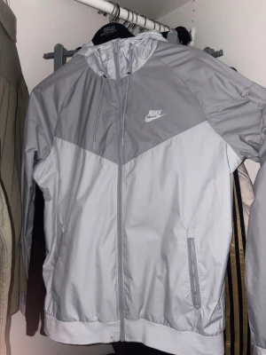Grå och vit vindjacka från Nike - Snygg tvåfärgad vindjacka från Nike i grått och vitt. Jackan har dragkedja framtill, justerbar huva med snören och två sidofickor med dragkedja. Tillverkad i lätt och ventilerande material med meshfoder för extra komfort.
