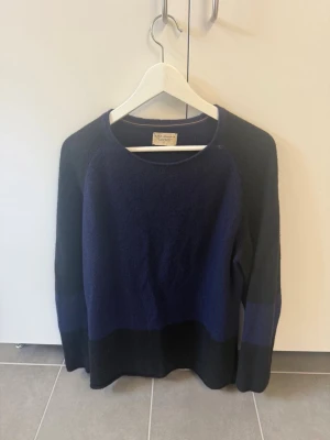 Mörkblå stickad tröja Nudie Jeans, stl M - Mörkblå stickad tröja från Nudie Jeans med rund halsringning och diskret färgblock i svart nedtill och på ärmarna. Tröjan har en avslappnad passform och är perfekt för lager på lager. Materialet ser mjukt och lite luddigt ut.