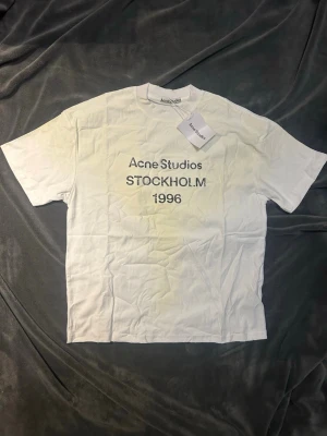 Vit M Acne Studios t-shirt  - Vit t-shirt från Acne Studios med trycket 'Acne Studios STOCKHOLM 1996' på bröstet. Klassisk passform och rund hals. Perfekt basplagg för en clean och stilren look. Köpt second hand därför det låga priset