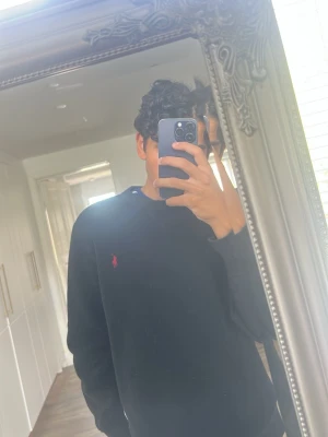 Svart sweatshirt från Polo Ralph Lauren - Svart sweatshirt från Polo Ralph Lauren med klassisk rund halsringning och röd broderad logga på bröstet. Tröjan har ribbade muddar vid ärmslut och nederkant, perfekt för en stilren och avslappnad look.