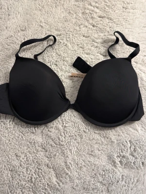 Svart push-up bh från Kappahl - En enkel och stilren svart push-up bh från Kappahl med släta kupor och justerbara axelband. Bh:n har bygel och vaddering för extra stöd och form. Perfekt under tighta toppar tack vare den sömlösa designen.