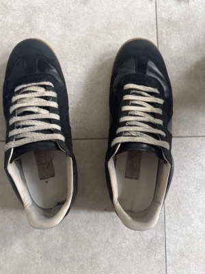Maison Margiela svarta sneakers - Svarta sneakers från Maison Margiela med beige snörning och vit insida. Ovandelen är i en mix av mocka och läder, och sulan är i klassisk gummifärg med tydlig logotyp undertill. Snygg retrostil som passar till det mesta.