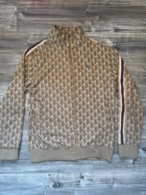 Beige Lacoste kofta - Jätte fin Lacoste kofta som sitter oerhört bra och verkligen avslutar en outfit till bästa. Vid fler intressen så är det bara att höra av er