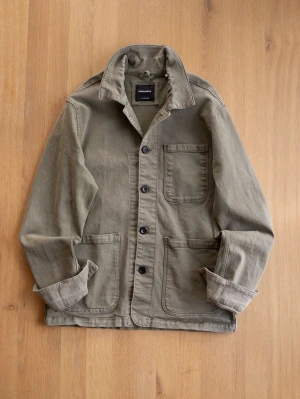 Green Chore Jacket, size XL - Snygg olivgrön overshirt i kraftig bomull med tre stora utanpåfickor och svarta knappar framtill. Klassisk krage och avslappnad passform, perfekt att slänga på över en t-shirt. Ärmarna kan vikas upp för en mer casual look.