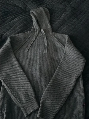 Grå stickad hoodie från Replay - Säljer en snygg grå stickad hoodie från Replay. Tröjan har ribbstickad struktur, långa ärmar och en huva med dragsko. Perfekt för dig som vill ha en stilren och bekväm stickad tröja till hösten eller vintern.