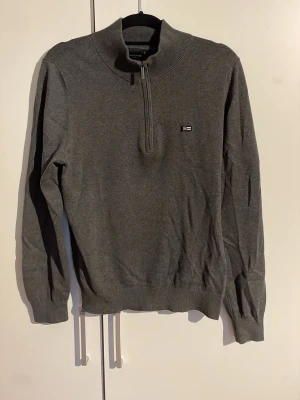 Grå halvzip tröja från lexington - Snygg grå långärmad tröja med halv dragkedja och ståkrage. Tröjan har ribbade muddar vid ärmslut och nederkant samt en liten broderad logga på bröstet. Perfekt för lager-på-lager och enkel att matcha med jeans eller chinos. Nypris 1000 kr
