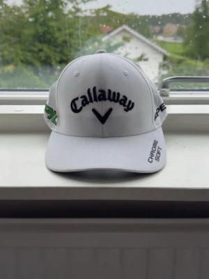 Vit Callaway keps med brodyr - Snygg vit keps från Callaway med broderade loggor i svart och grönt. Framtill står det 'Callaway' och 'Chrome Soft', på sidan 'EPIC' och baktill 'Odyssey'. Justerbar rem bak för perfekt passform. Perfekt för golf eller streetwear.