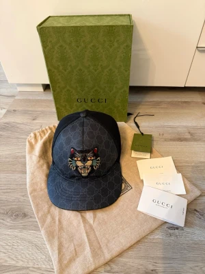 Gucci keps - Snygg svart keps från Gucci med klassiskt GG-mönster och broderad tiger framtill. Kepsen har mesh på sidorna och justerbar rem bak. Tillverkad i bomull och polyester, perfekt för dig som vill sticka ut med en exklusiv accessoar. ÄKTA! Allt på bilden följer med!! Vid frågor eller funderingar så är det bara att höra av sig!!