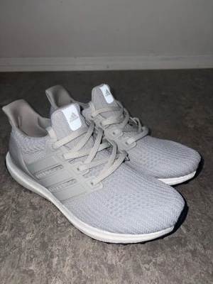 Grå Adidas Ultraboost sneakers - Snygga grå Adidas Ultraboost sneakers med stickad ovandel i mesh och klassiska tre ränder på sidan. Skorna har vita sulor med Boost-dämpning och gråa skosnören. Perfekta för dig som gillar stilrena och bekväma sneakers.