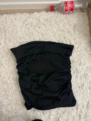Svart bandeau tubtopp - Säljer en svart bandeau tubtopp med enkel och stilren design. Toppen är axelbandslös och har lätt rynkade sidor för en snygg passform. Perfekt att matcha med jeans eller kjol för en trendig look.